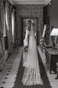 Load image into Gallery viewer, Woman in a wedding dress with a long veil standing in a room with patterned curtains and decor.