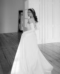Load image into Gallery viewer, Woman in a white wedding dress standing in a room with wooden floors and white walls.