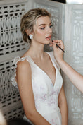 Load image into Gallery viewer, Bride in a white lace dress getting makeup applied in a decorative setting.