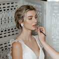 Load image into Gallery viewer, Bride in a white lace dress getting makeup applied in a decorative setting.