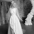 Load image into Gallery viewer, Black and white photo of a woman in a wedding dress with a floral arch and brick wall background.