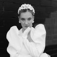 Load image into Gallery viewer, Woman in a white dress with puffed sleeves and floral headpiece against a dark brick wall.