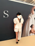 Load image into Gallery viewer, Woman in a white outfit standing in front of a branded backdrop with a large 'S' logo.