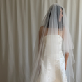Charger l'image dans la visionneuse de la galerie, Bride in a white wedding dress with a long veil standing against a white curtain.