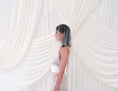 Load image into Gallery viewer, A woman in profile wearing a sheer crystal bridal veil and a white lace wedding dress, standing in front of draped white fabric.