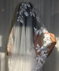 Charger l'image dans la visionneuse de la galerie, A woman wearing a long white bridal veil with floral lace appliqué and a matching sleeveless wedding dress, with a white curtain background.