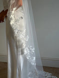 Load image into Gallery viewer, A close-up of a white satin wedding dress with a long sheer tulle veil featuring intricate white floral lace applique, worn by a model standing indoors against a neutral gray wall.