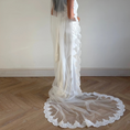 Bild in Galerie-Betrachter laden, A woman wearing a white wedding dress and long sheer lace veil stands in profile in a room with a light wall and wood floor. The dress has a fitted bodice, cap sleeves, and a dramatic circular lace-trimmed train.