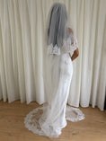 Load image into Gallery viewer, Person wearing a white lace wedding dress with a long veil in front of white curtains.