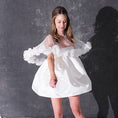 Load image into Gallery viewer, A woman modeling a short white dress with a 3d flower tulle bridal cape standing against a dark textured studio wall.