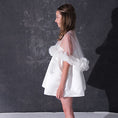 Load image into Gallery viewer, A woman modeling a short white dress with a 3d flower tulle bridal cape standing against a dark textured studio wall.