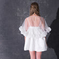 Load image into Gallery viewer, A woman modeling a short white dress with a 3d flower tulle bridal cape standing against a dark textured studio wall.