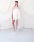Load image into Gallery viewer, A woman wearing a sheer white bridal veil with delicate bow details and a short white slip dress, standing in front of draped white fabric.