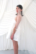 Load image into Gallery viewer, A woman wearing a sheer white bridal veil with delicate bow details and a short white slip dress, standing in front of draped white fabric.