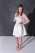 Load image into Gallery viewer, A woman modeling a short white dress with a pearl tulle bridal cape standing against a dark textured studio wall.