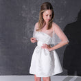 Load image into Gallery viewer, A woman modeling a short white dress with a pearl tulle bridal cape standing against a dark textured studio wall.