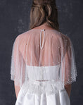Load image into Gallery viewer, A woman modeling a short white dress with a pearl tulle bridal cape standing against a dark textured studio wall.