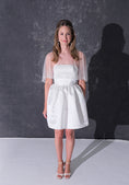 Load image into Gallery viewer, A woman modeling a short white dress with a pearl tulle bridal cape standing against a dark textured studio wall.