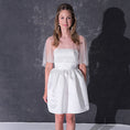 Load image into Gallery viewer, A woman modeling a short white dress with a pearl tulle bridal cape standing against a dark textured studio wall.