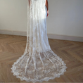 Load image into Gallery viewer, A woman viewed from behind wearing a long white lace bridal veil over her head and shoulders, standing on a light wood herringbone floor against a plain off-white wall.