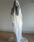 Charger l'image dans la visionneuse de la galerie, A woman wearing a long white lace bridal veil over her head and shoulders, paired with a white wedding dress, standing on a light wood herringbone floor against a plain off-white wall.