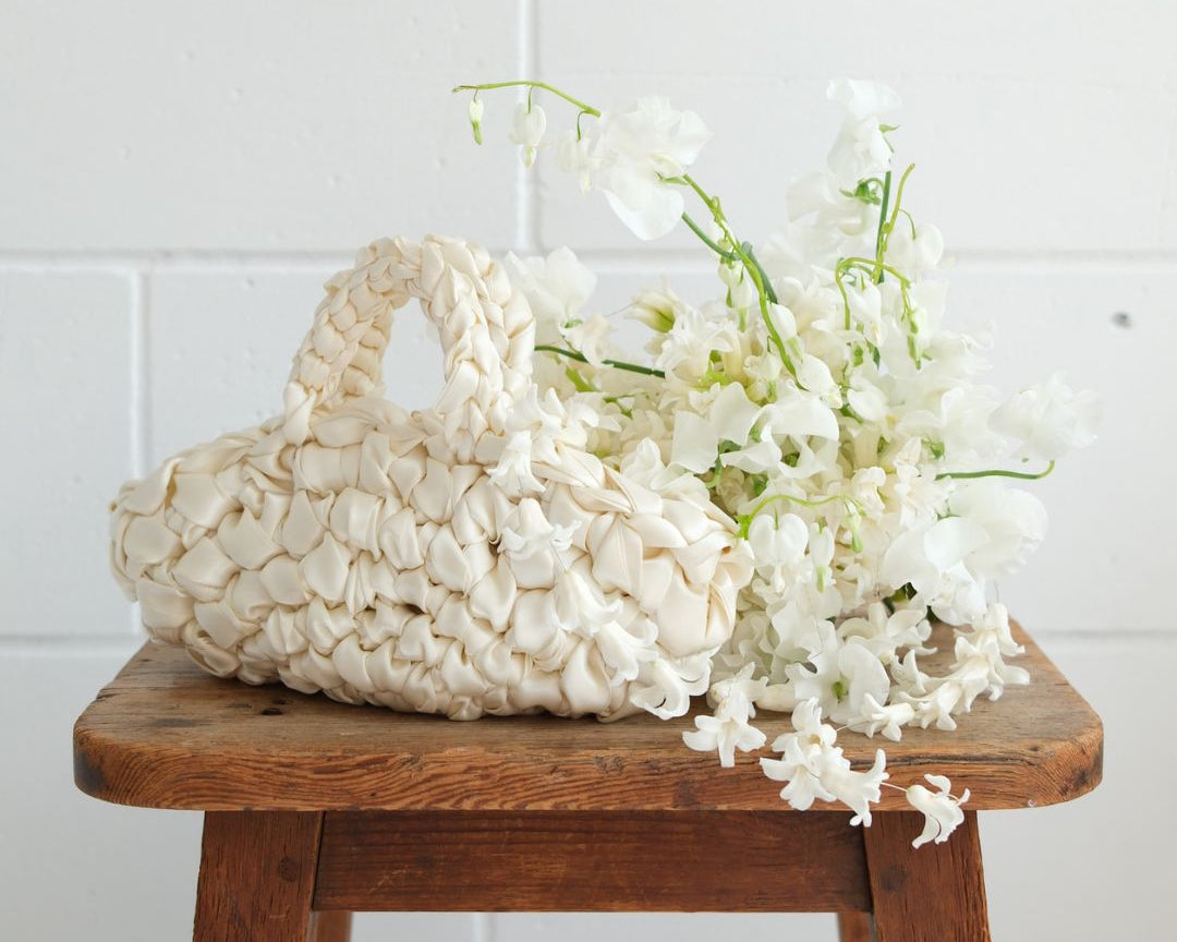 White woven bag with white flowers on a wooden stool against a white tiled wall.