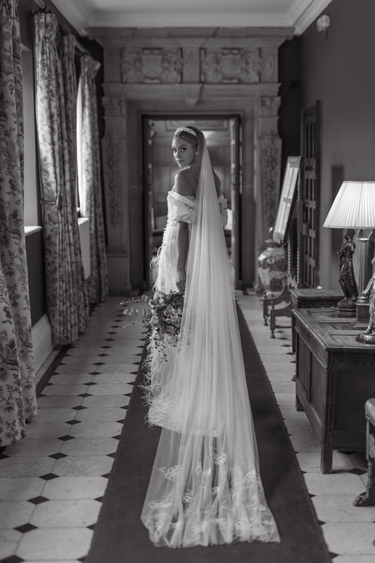 Woman in a wedding dress with a long veil standing in a room with patterned curtains and decor.