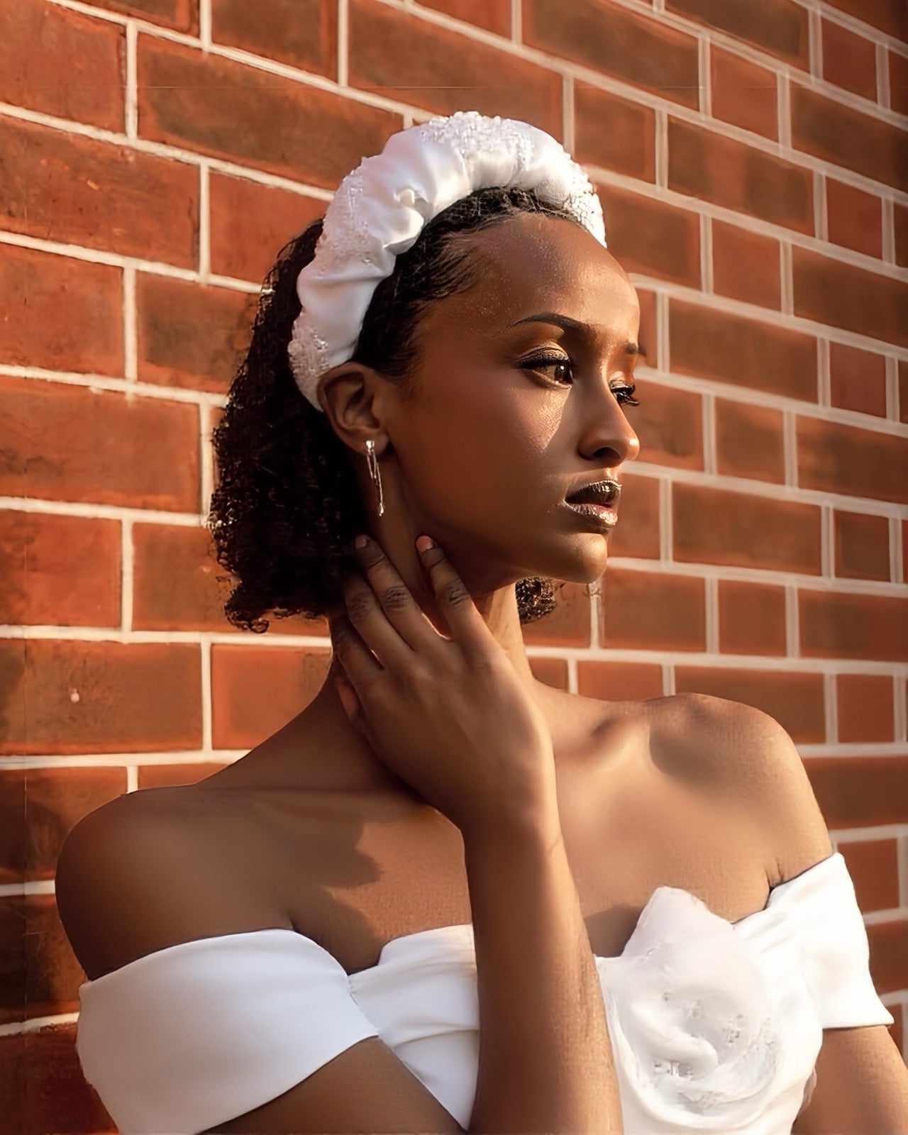 Woman wearing a white off-shoulder top and bridal headband against a brick wall.