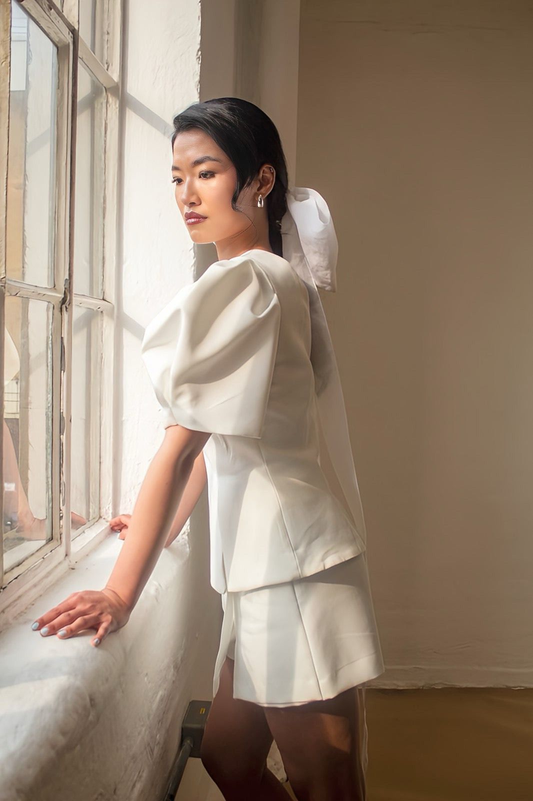 Woman in a white outfit standing by a window with soft natural light.