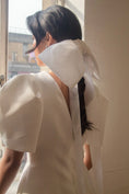 Load image into Gallery viewer, Person wearing a white dress with a large bridal bow at the neck, standing by a window.
