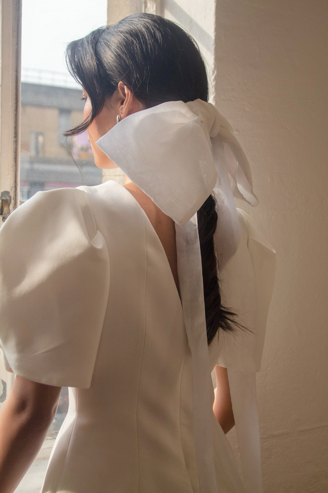 Person wearing a white dress with a large bridal bow at the neck, standing by a window.