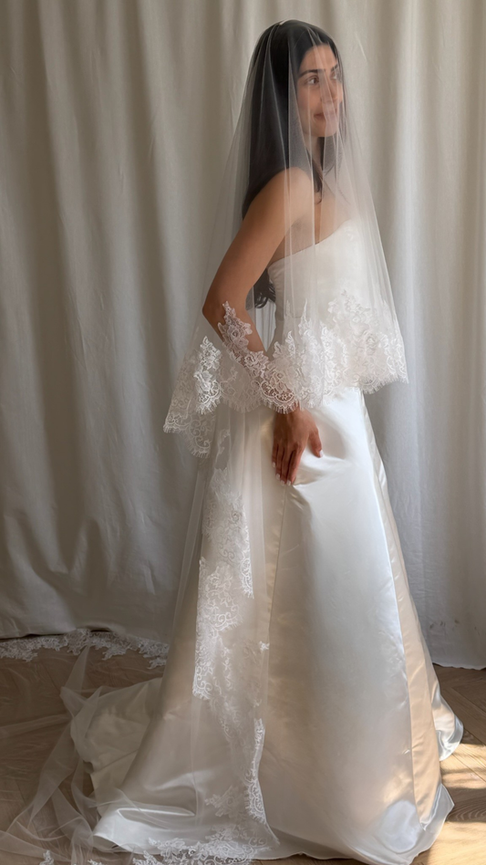 Bride wearing a white wedding dress with a lace veil against a white curtain background.