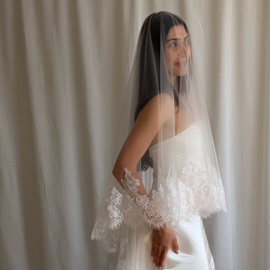 Bride wearing a white wedding dress with a lace veil against a white curtain background.