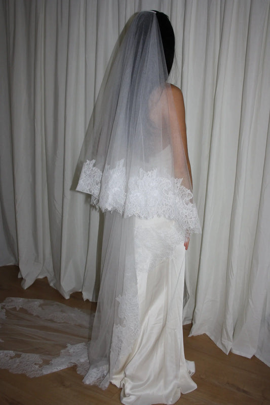 Bride wearing a white wedding dress with a lace veil against a white curtain background.