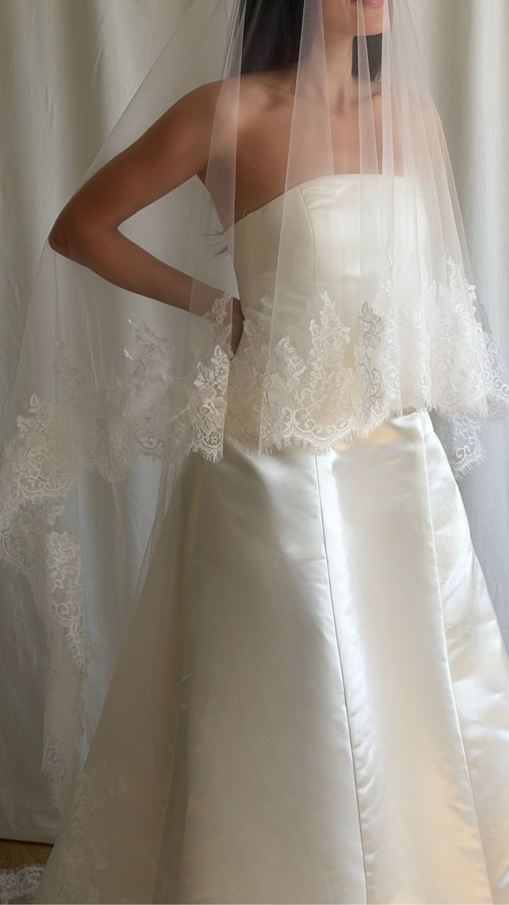 Bride wearing a white wedding dress with a lace veil against a white curtain background.