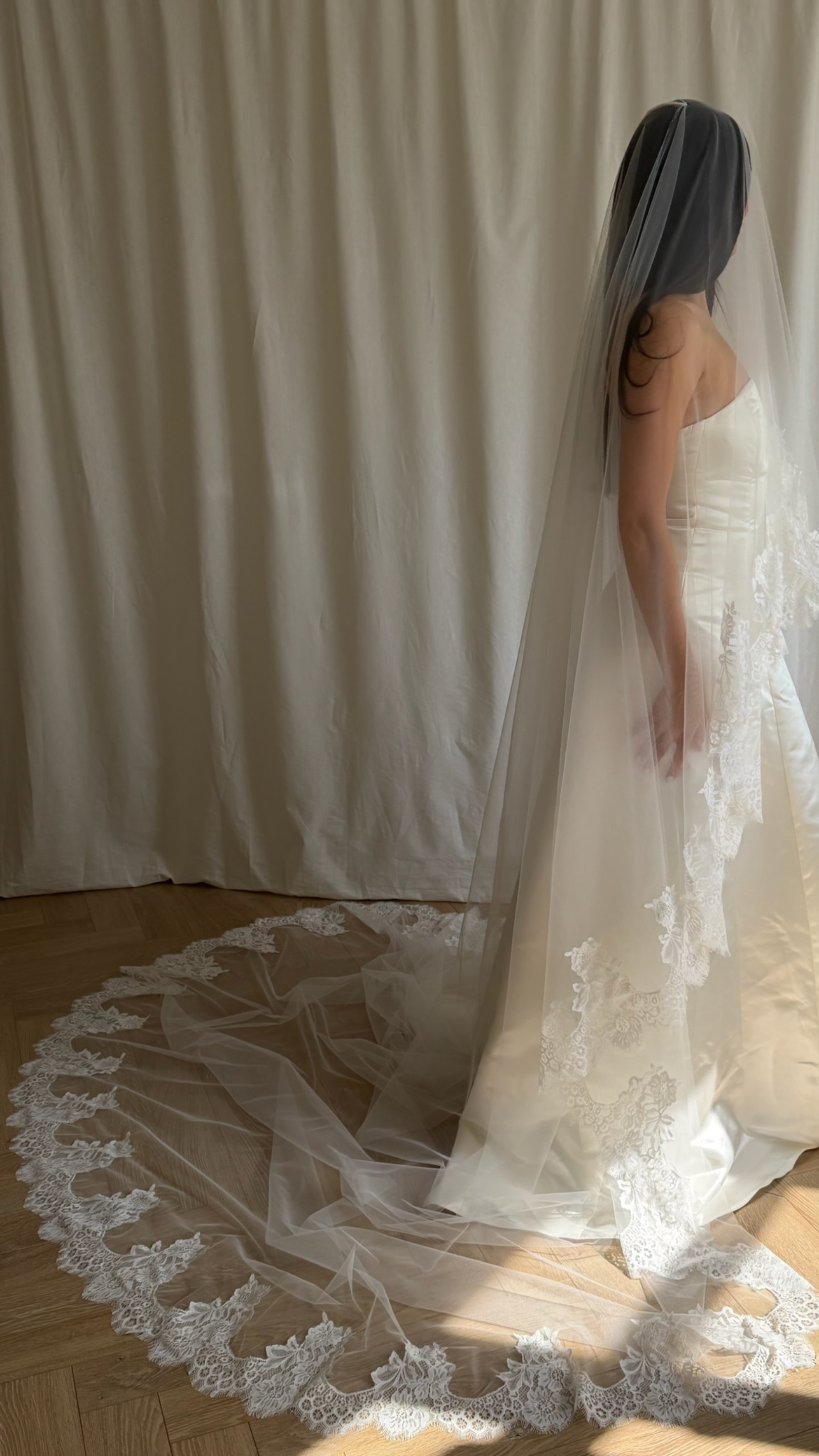 Bride wearing a white wedding dress with a lace veil against a white curtain background.