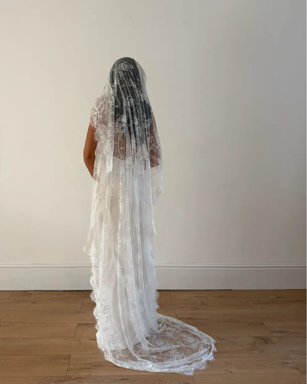 Person wearing a white wedding dress with a long lace veil against a plain background
