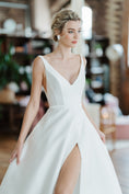 Load image into Gallery viewer, Woman wearing a white wedding dress with a slit in a blurred indoor setting
