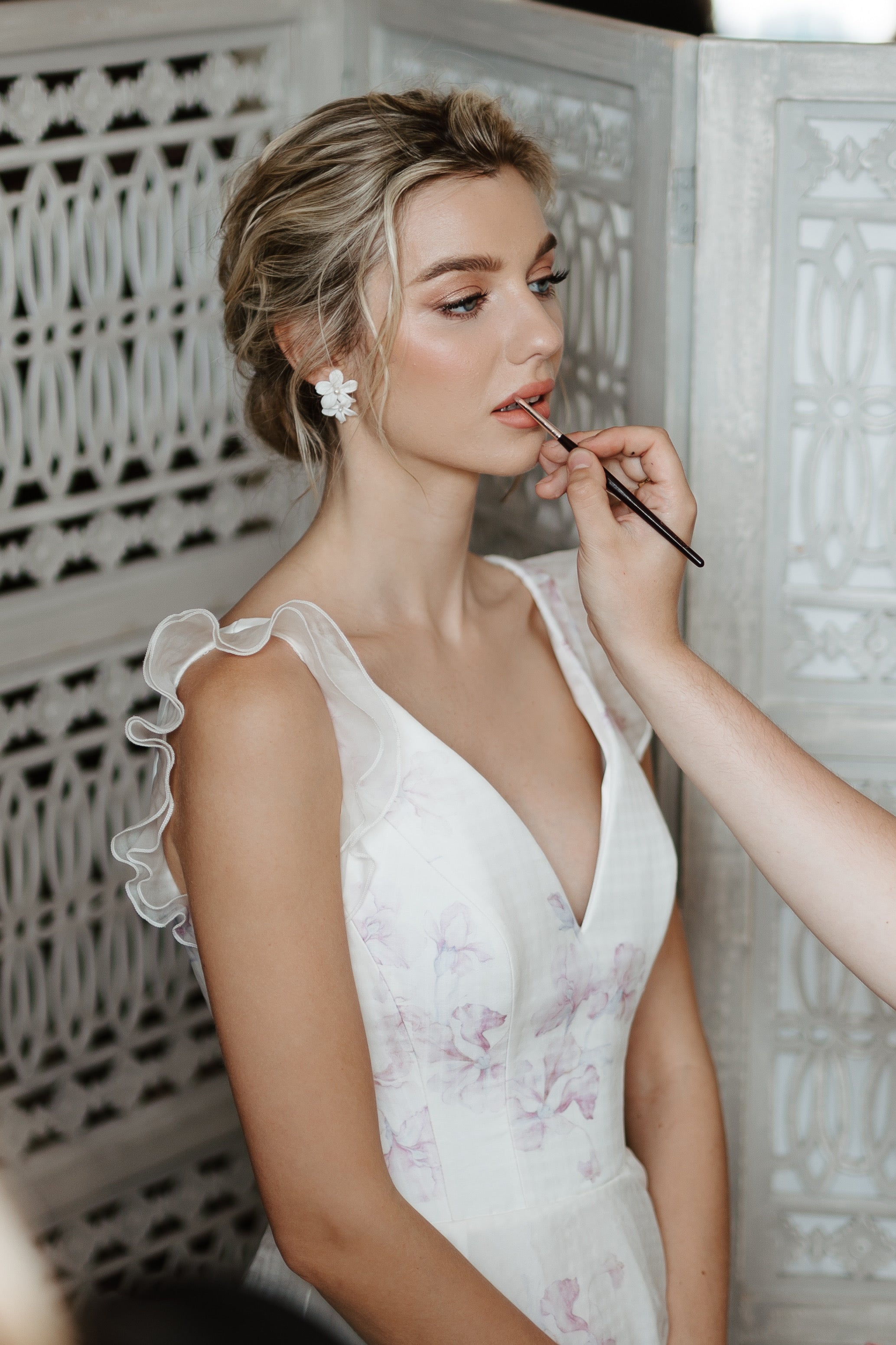 Bride in a white lace dress getting makeup applied in a decorative setting.
