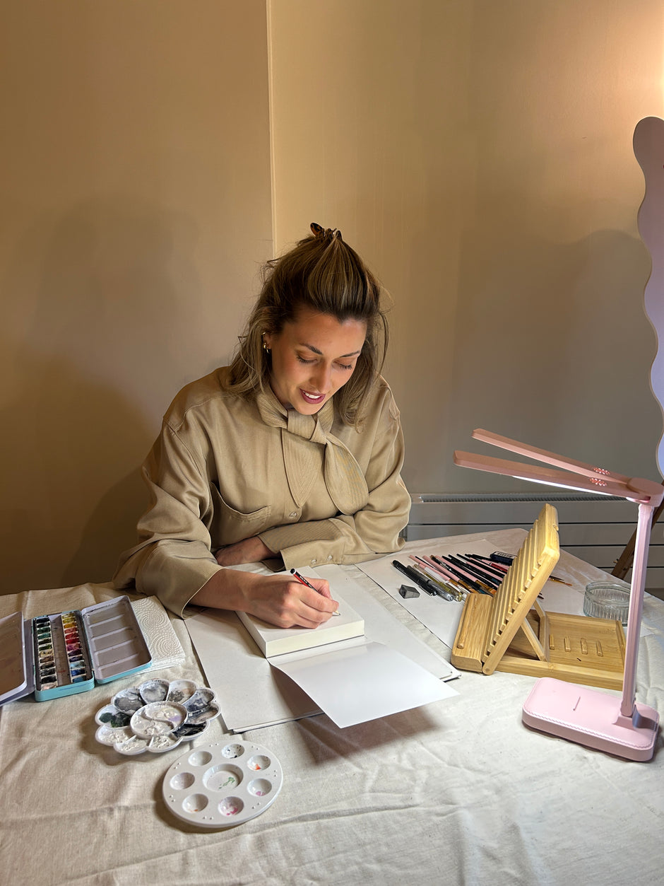 A woman in a beige jacket is seen from above writing in a notebook at a desk covered with art supplies, including a pink lamp, wooden stand, paint palette, and colored pencils.