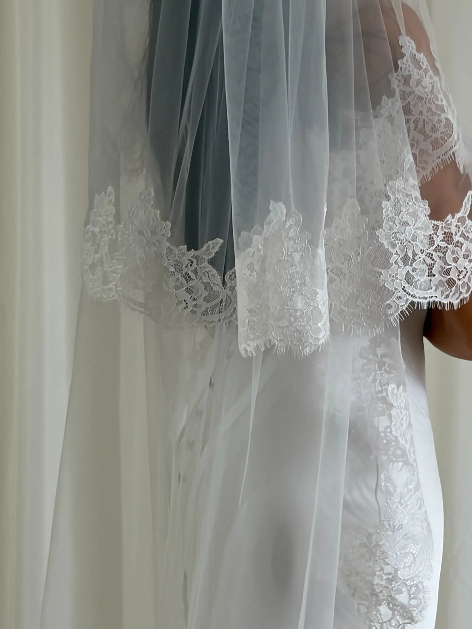 close uo of white wedding dress with a long lace veil in front of a white curtain.