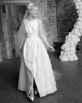 Load image into Gallery viewer, Black and white photo of a woman in a wedding dress with a floral arch and brick wall background.
