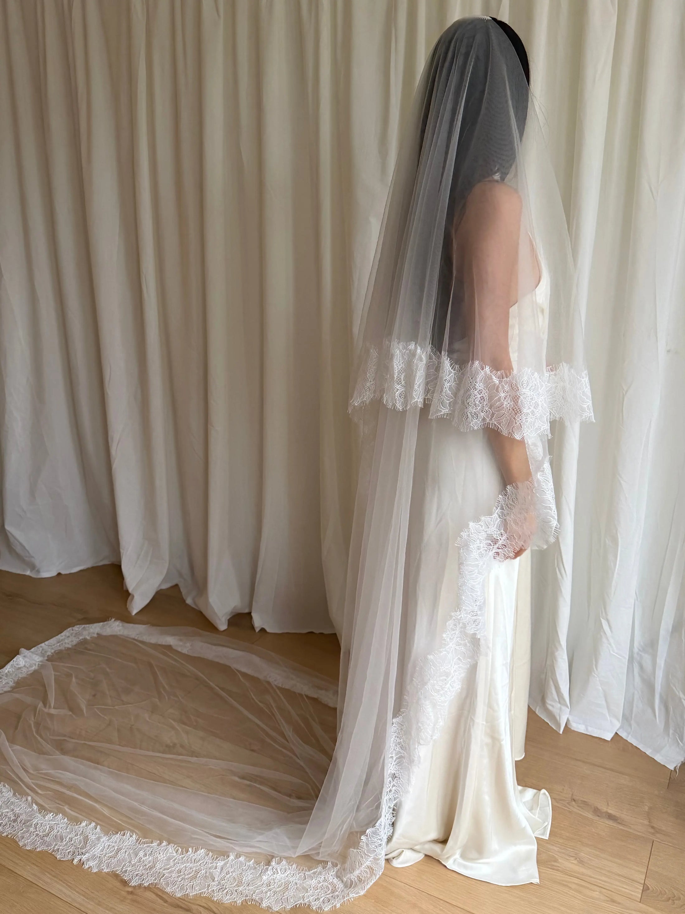 Bride wearing a white wedding dress with a lace veil against a white curtain background.