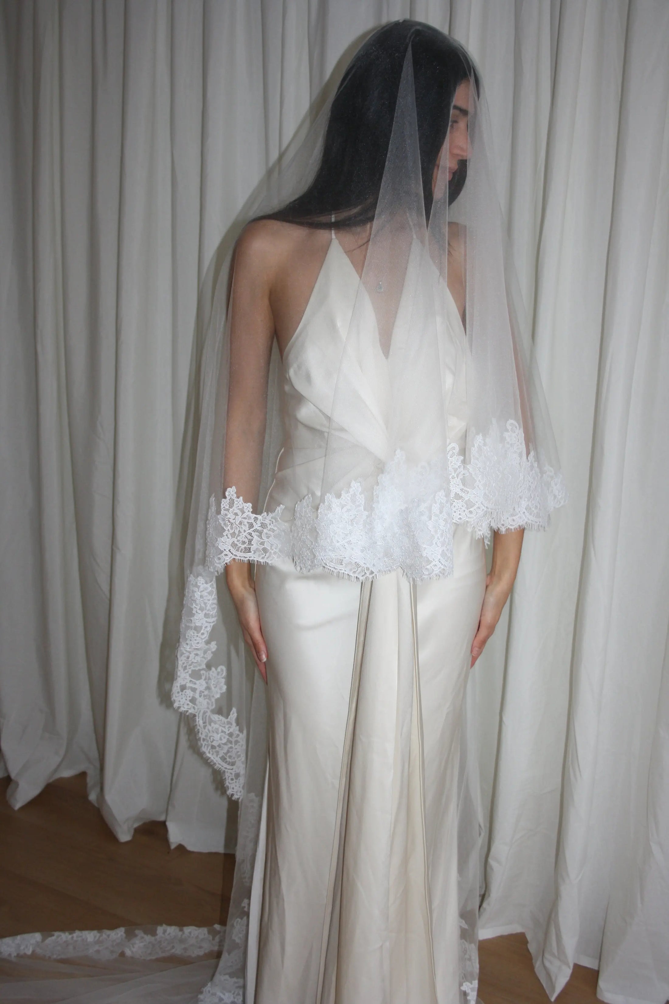 person in a wedding dress with a long lace trim veil against a curtain backdrop.