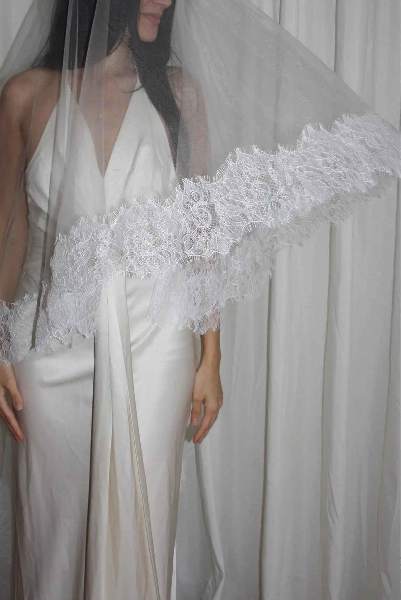 Bride wearing a white wedding dress with a lace veil against a white curtain background.