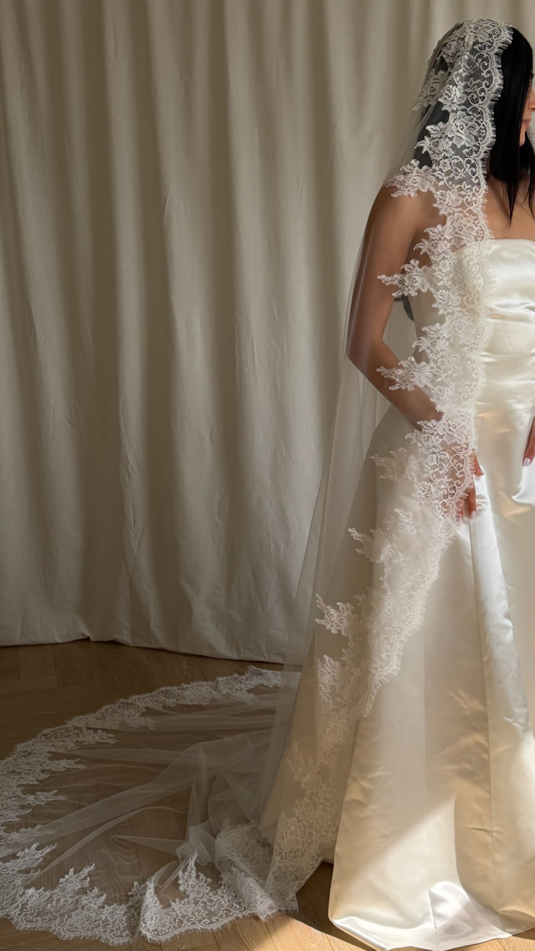 Bride wearing a white wedding dress with a lace veil against a white curtain background.