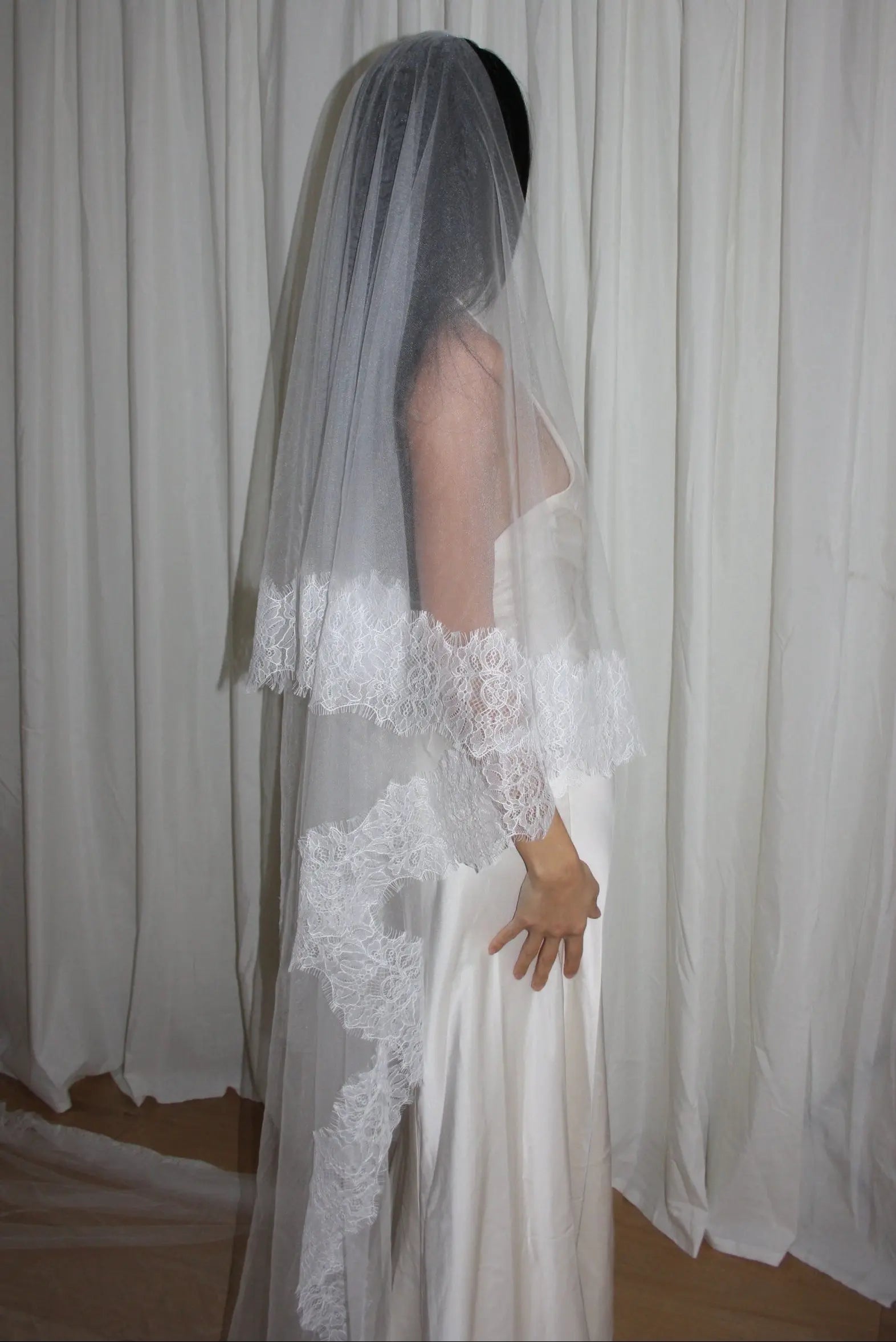 Bride wearing a white wedding dress with a lace veil against a white curtain background.