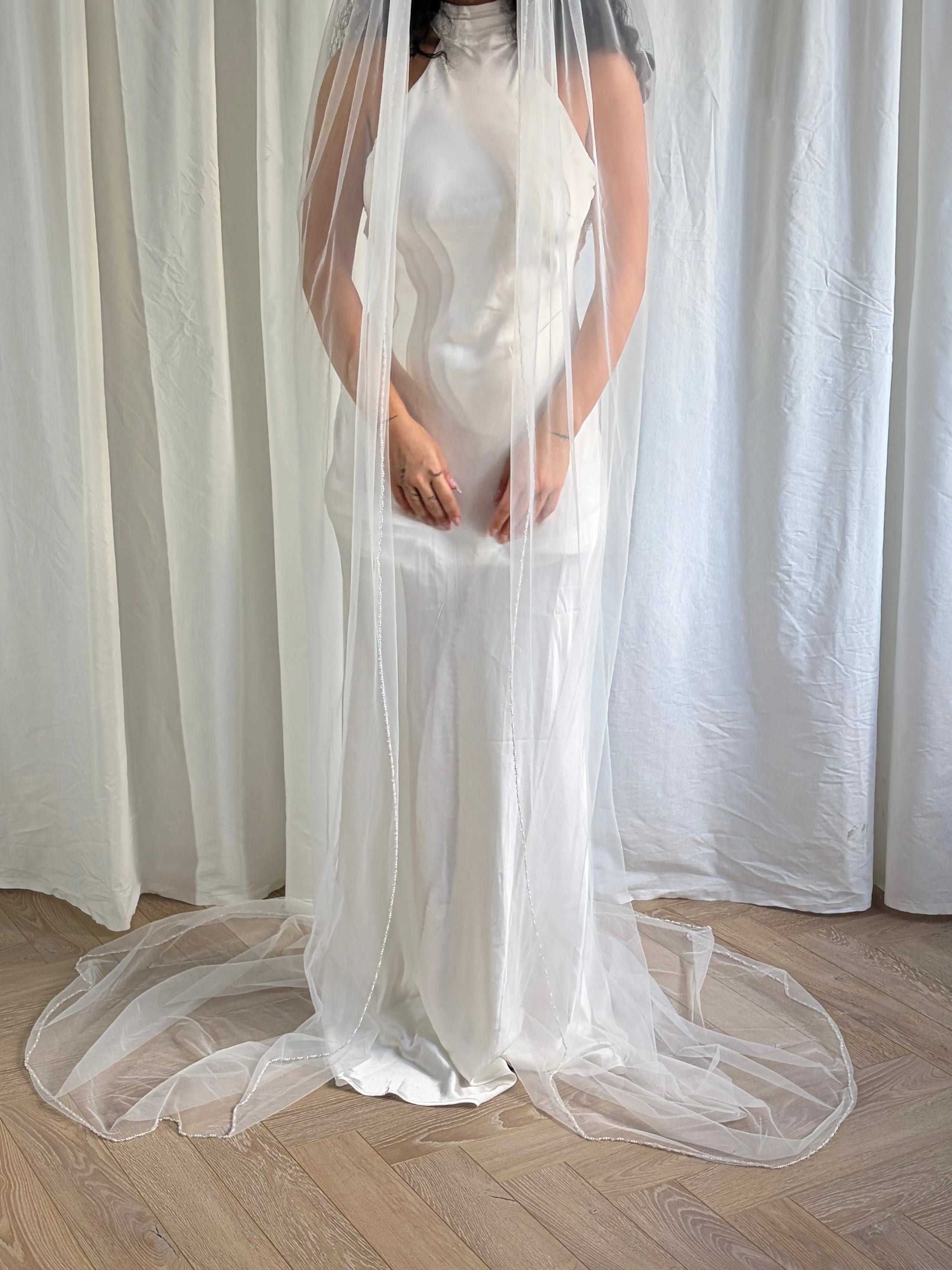 Person wearing a white wedding dress with a long pearl veil against a plain background