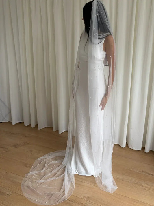 Person wearing a white wedding dress with a long pearl veil against a plain background