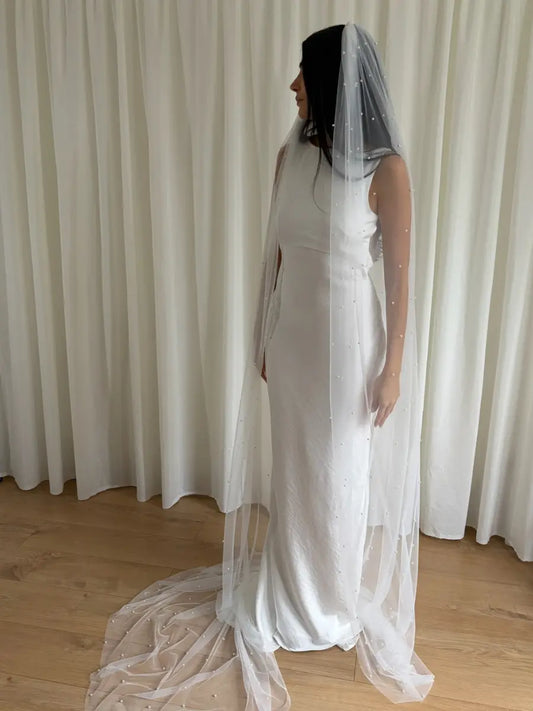 Person wearing a white wedding dress with a long pearl veil against a plain background